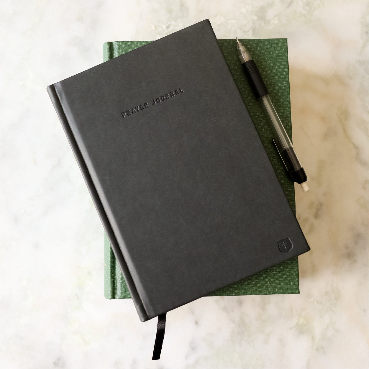 Black prayer journal with a pen on a green book against a marble background
