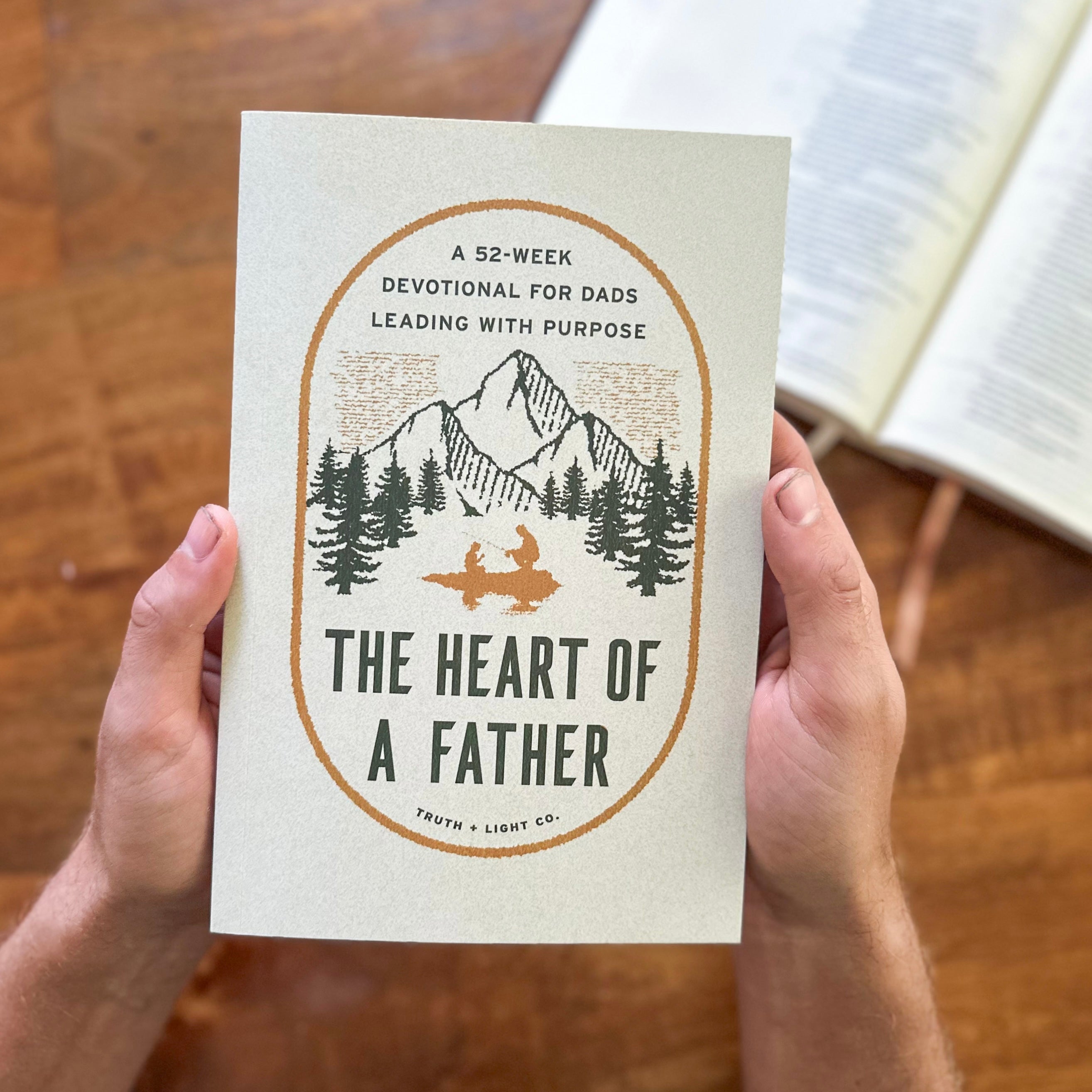 Book titled 'The Heart of a Father' held by hands on a wooden surface with an open book in the background.